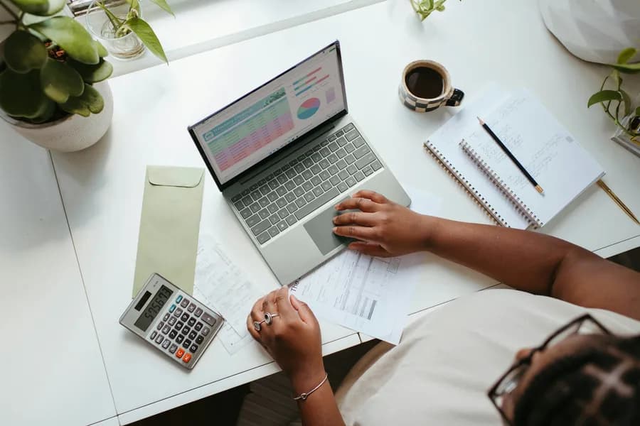 Hands working on a laptop beside a notebook, calculator and cup of tea